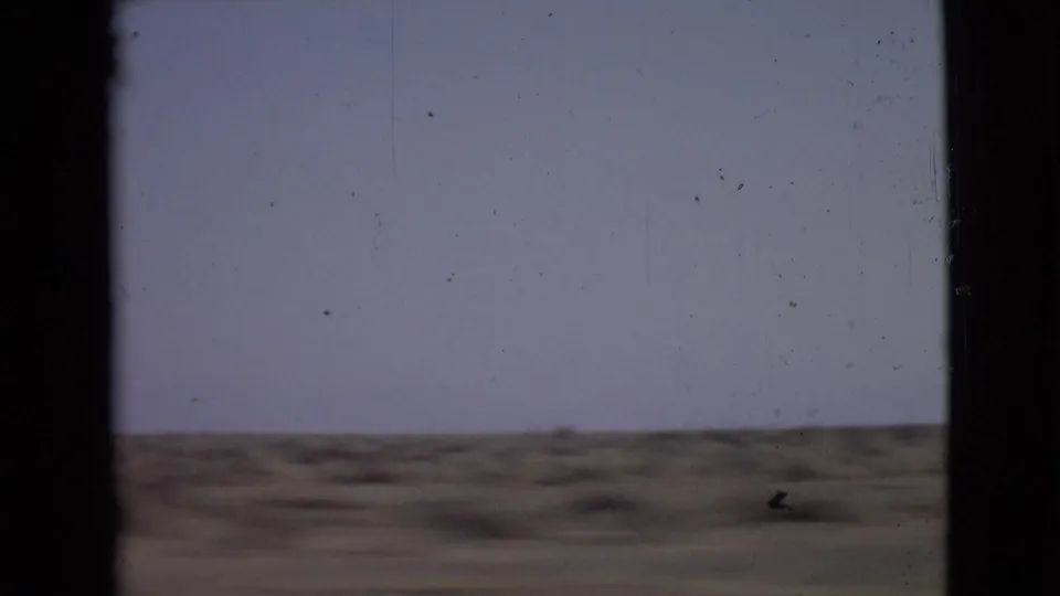 1968 home-movie frame from a moving car, showing flat arid ground, a distant low horizon, and visible 8mm dust and scratches.