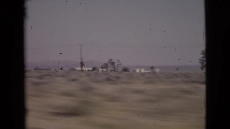 1968 8mm view from a moving car across arid San Diego County terrain with scattered trees, utility poles, and low distant buildings.