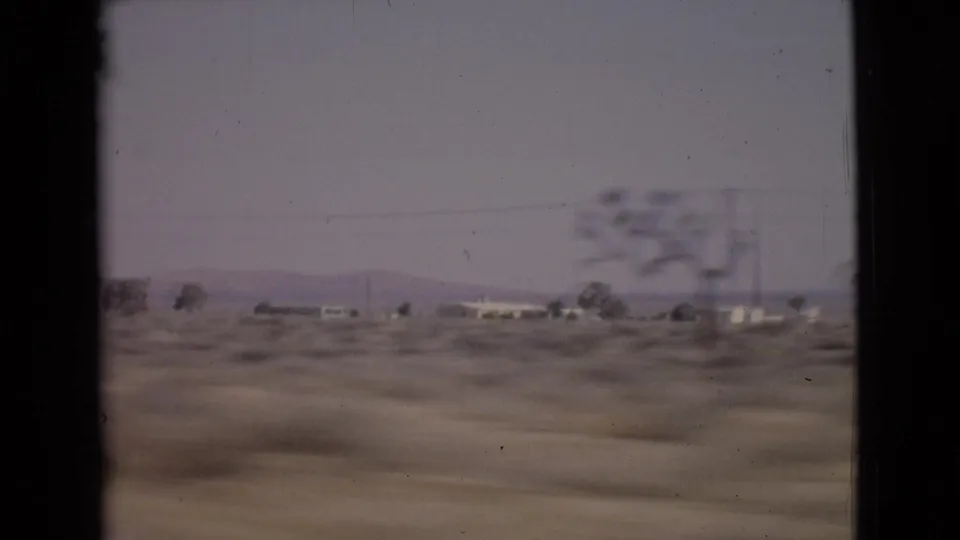 Moving 8mm car-window view of San Diego County scrubland with low buildings and sparse trees under a pale hazy sky.