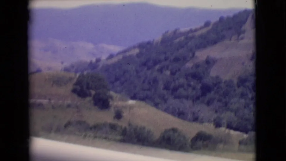1968 San Diego County 8mm frame showing rolling hills, wooded patches, and distant ridgelines under a hazy sky.