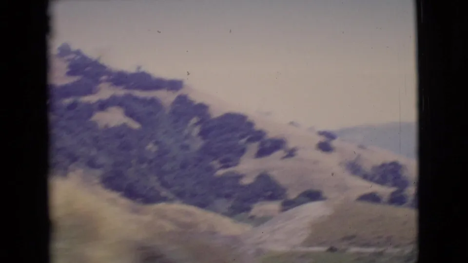 Soft 8mm view of San Diego County hillside terrain with scattered trees and pale slopes, likely filmed from a moving vehicle.