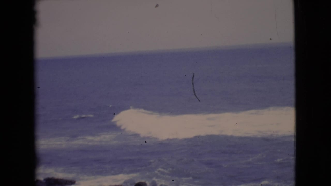 White ocean wave breaking beyond dark rocks with black film borders.