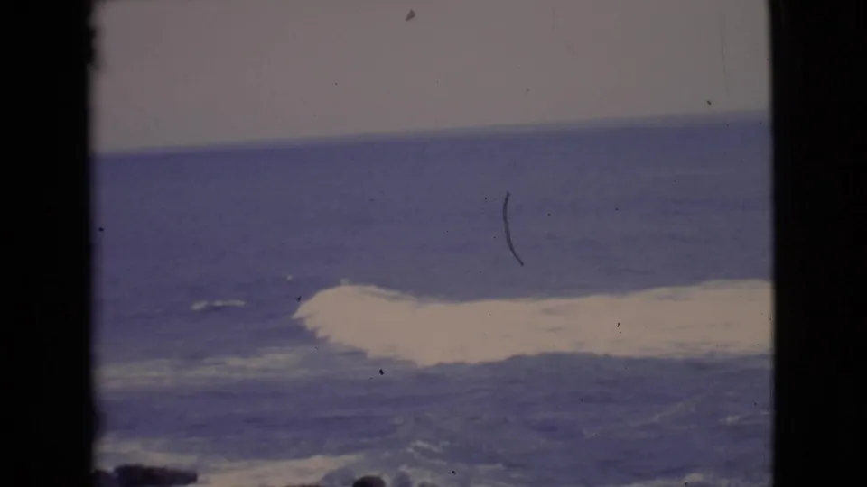 1968 8mm San Diego County coastal view of a white wave breaking offshore beyond dark rocks and a blue horizon.