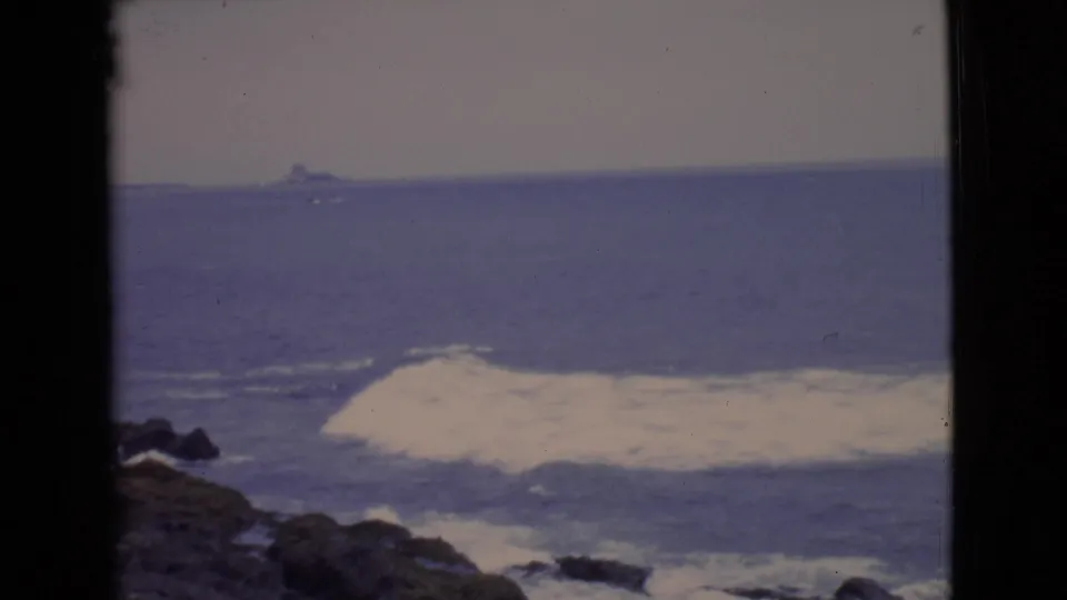1968 San Diego County 8mm frame with a foamy surf line, dark shoreline rocks, and a faint offshore form on the horizon.