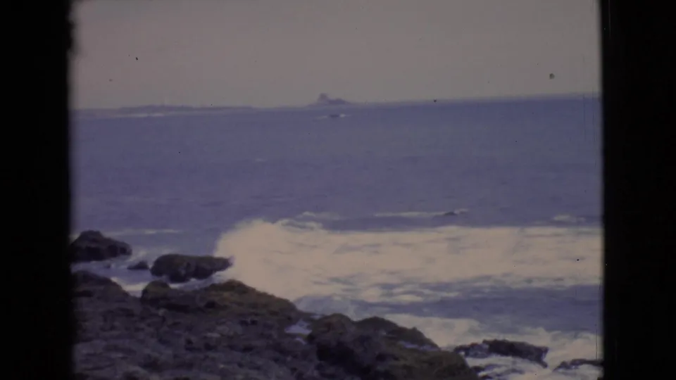 1968 8mm coastal frame from San Diego County showing white foam against dark rocks and a faint distant form offshore.