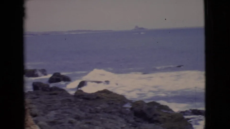 1968 8mm home-movie view of San Diego County surf breaking over dark coastal rocks beneath a hazy horizon.