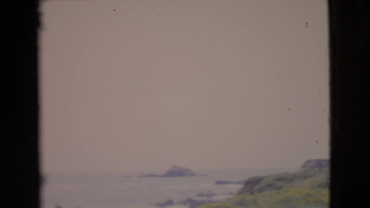 1968 San Diego County 8mm frame showing a hazy Pacific coast, distant sea stack, rocky slope, and dark vertical frame edges.