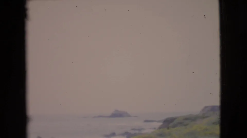 1968 San Diego County 8mm frame showing a hazy Pacific coast, distant sea stack, rocky slope, and dark vertical frame edges.
