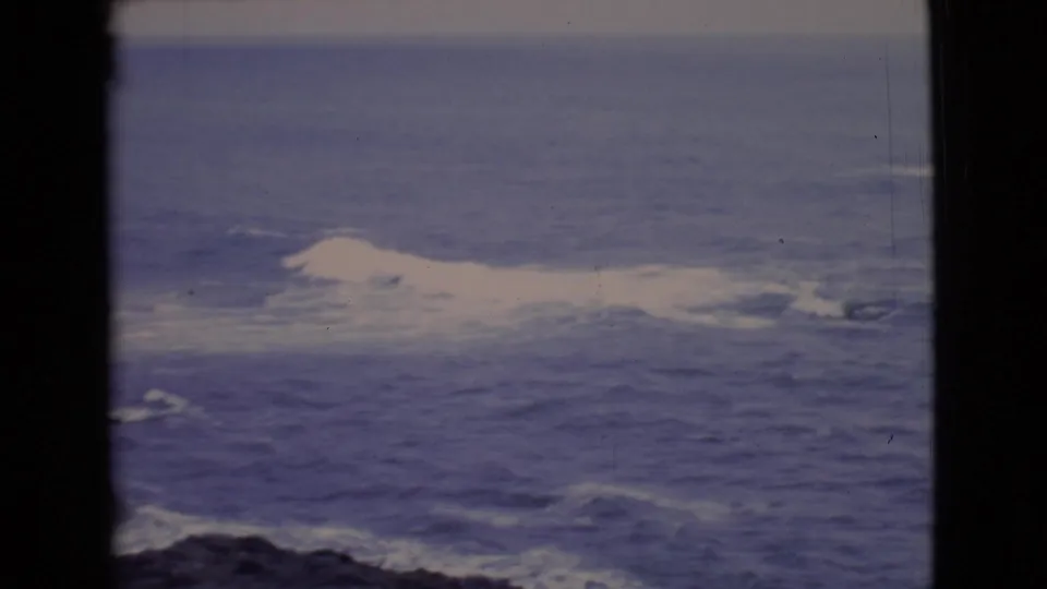 1968 San Diego County 8mm view of turbulent blue ocean and white surf near dark coastal rocks, seen through heavy frame edges.