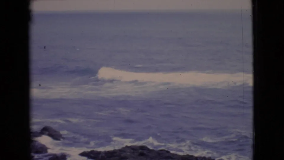 1968 San Diego County coastal frame with a white wave crest, blue Pacific water, dark foreground rocks, and soft 8mm color.