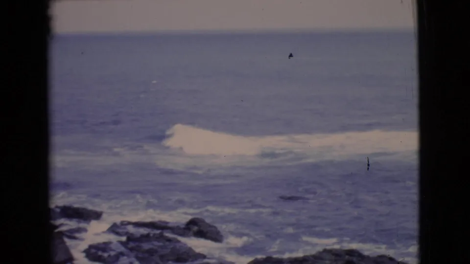 1968 San Diego County 8mm frame of open Pacific water, foamy surf, dark coastal rocks, and a hazy horizon line.
