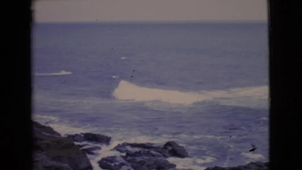 1968 San Diego coastal view with blue-gray ocean, breaking waves, rocks, and dark window framing at the edge of the frame.