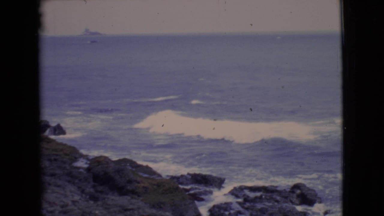 San Diego County shoreline in 1968, with white foam rolling toward rocks and a distant shape near the horizon.