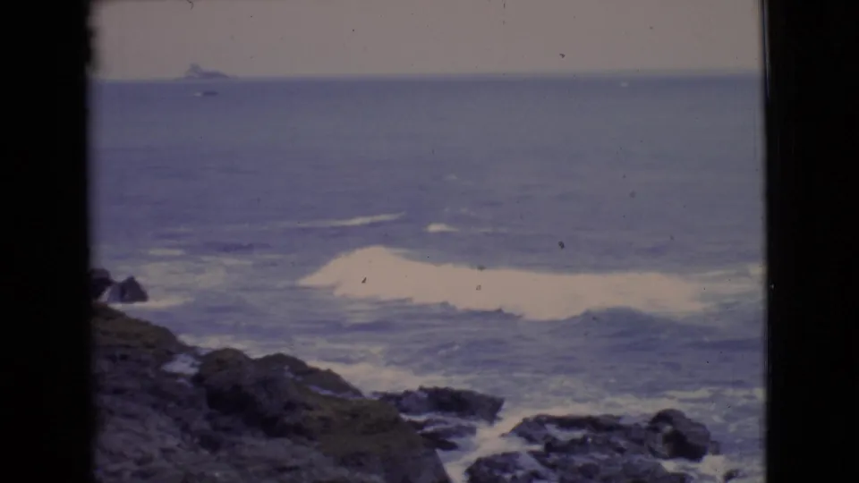 San Diego County shoreline in 1968, with white foam rolling toward rocks and a distant shape near the horizon.