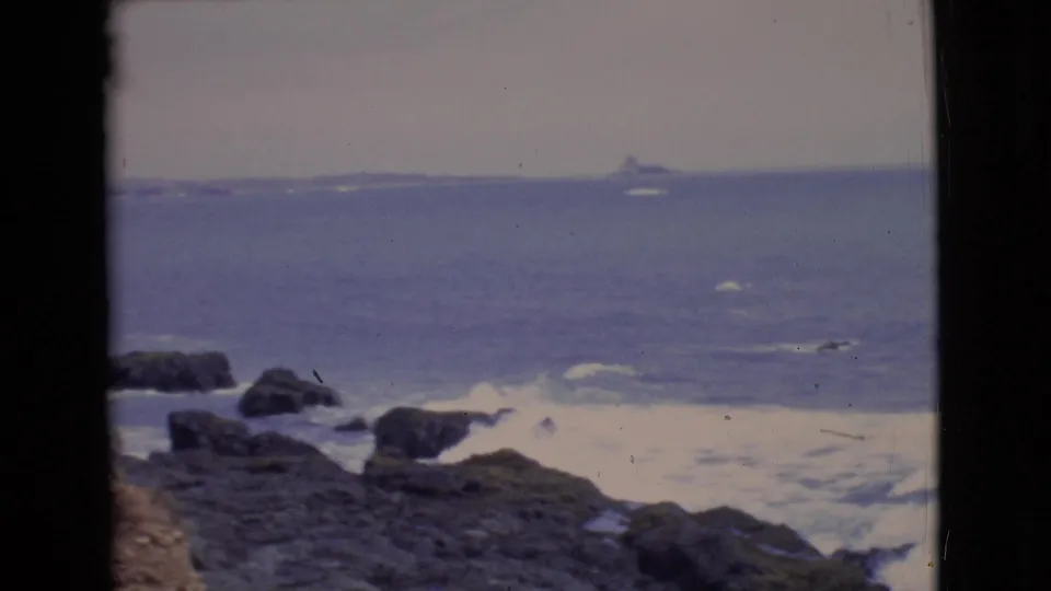 1968 home-movie frame of San Diego surf breaking over coastal rocks, with a faint distant landform or vessel on the horizon.