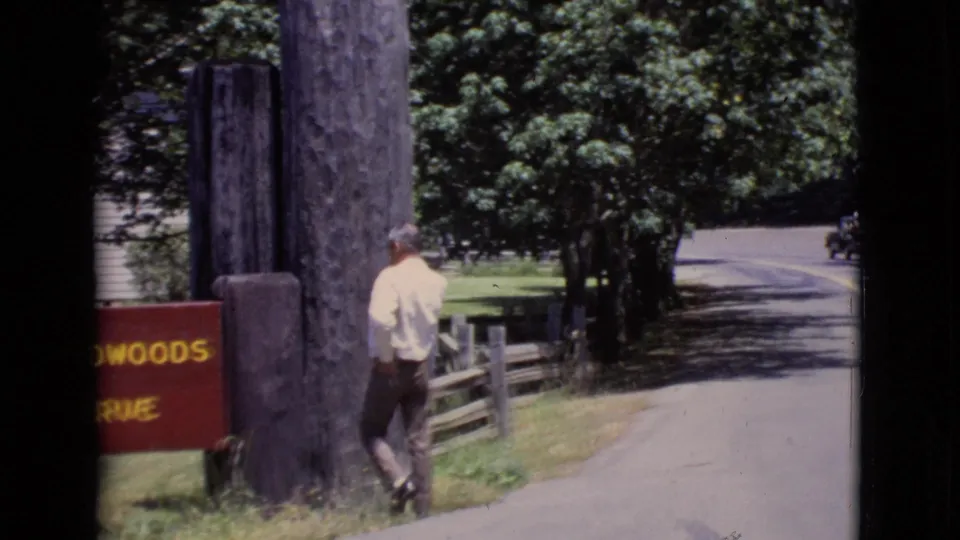 A man walks near a partially visible red state reserve sign in a wooded California park setting, with road and fence behind him.
