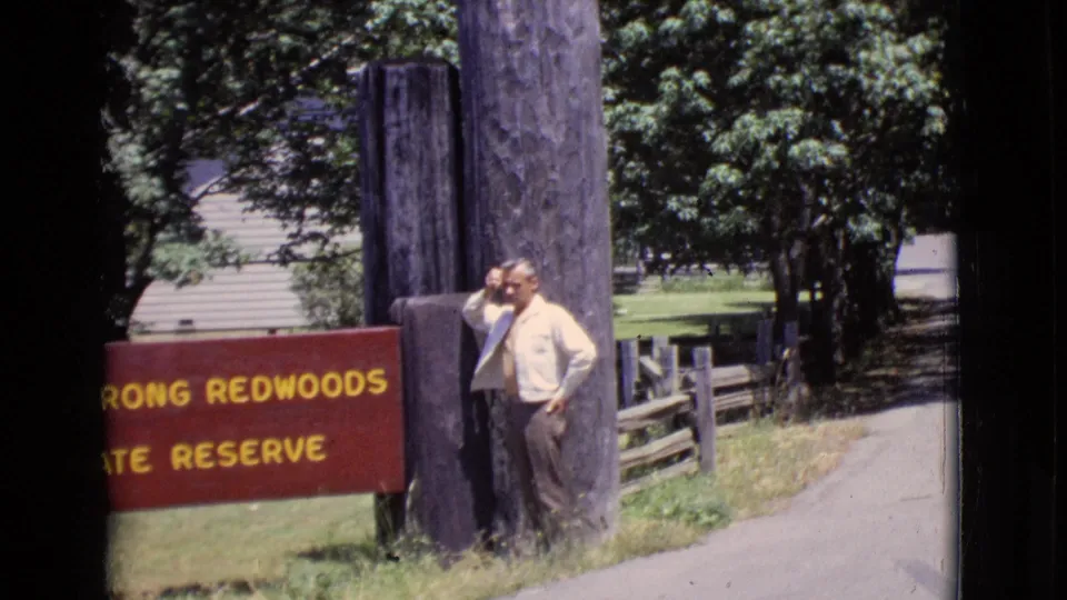 In 1968 California footage, a man stands by a red sign reading in part Strong Redwoods State Reserve near trees and a paved road.