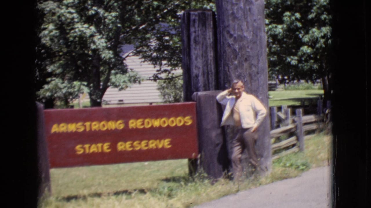 1968 home-movie frame of a man posing beside the Armstrong Redwoods State Reserve sign in California, with trees and a paved path nearby.