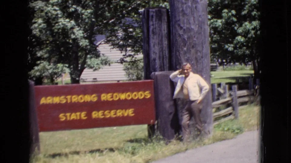 1968 home-movie frame of a man posing beside the Armstrong Redwoods State Reserve sign in California, with trees and a paved path nearby.