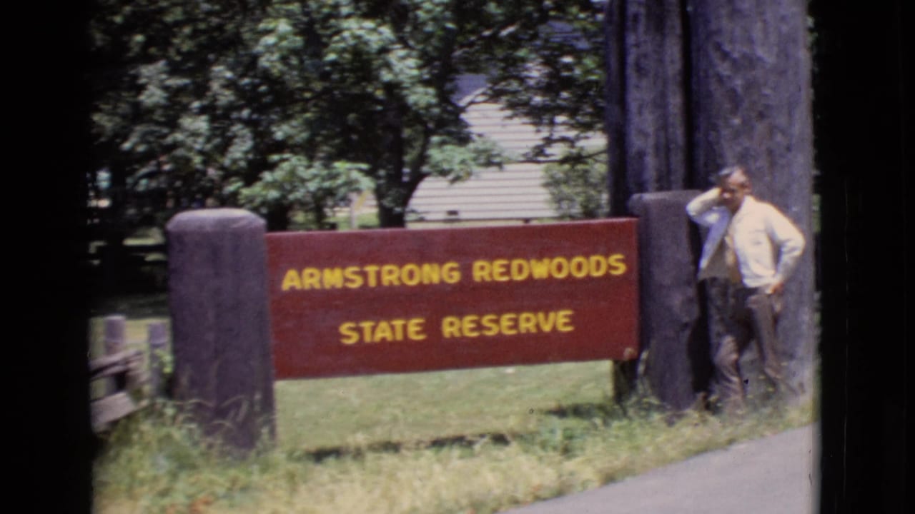 The Armstrong Redwoods State Reserve sign fills the frame as a man stands casually at the right edge near a large timber post.