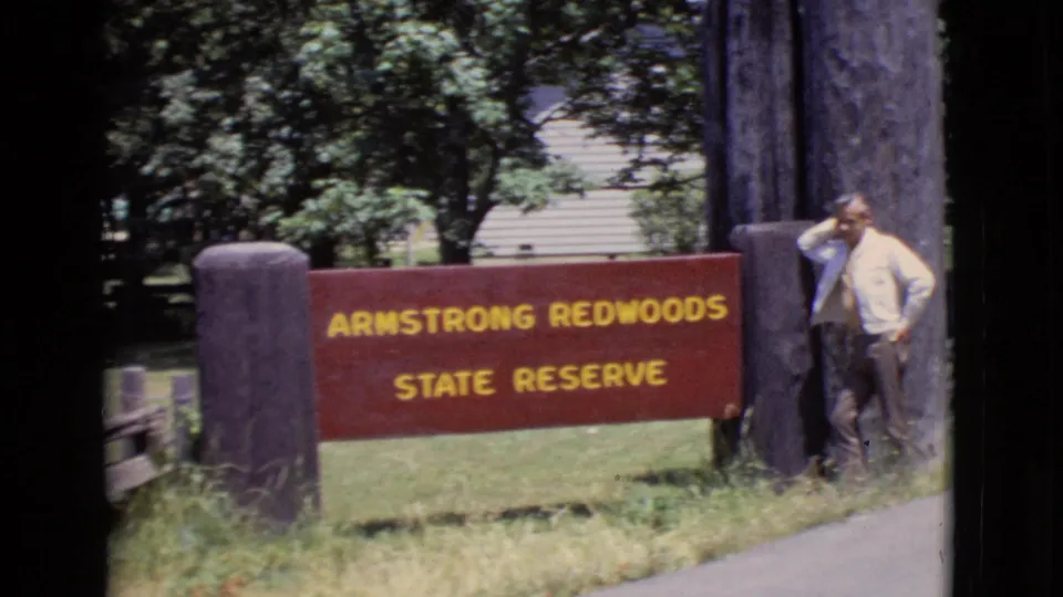 The Armstrong Redwoods State Reserve sign fills the frame as a man stands casually at the right edge near a large timber post.