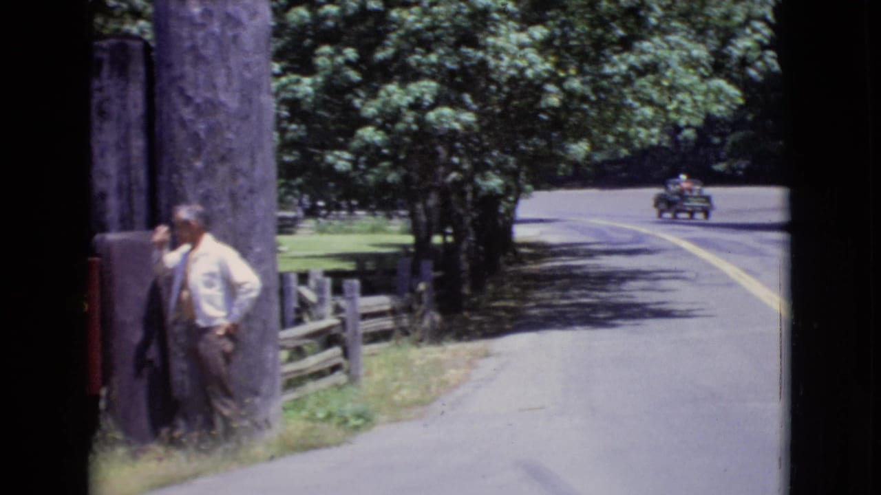1968 San Diego County frame with the man near a wooden post, roadside fence and trees, and a small vintage car moving along the curving road.