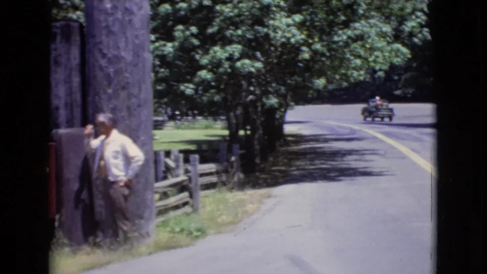 1968 San Diego County frame with the man near a wooden post, roadside fence and trees, and a small vintage car moving along the curving road.