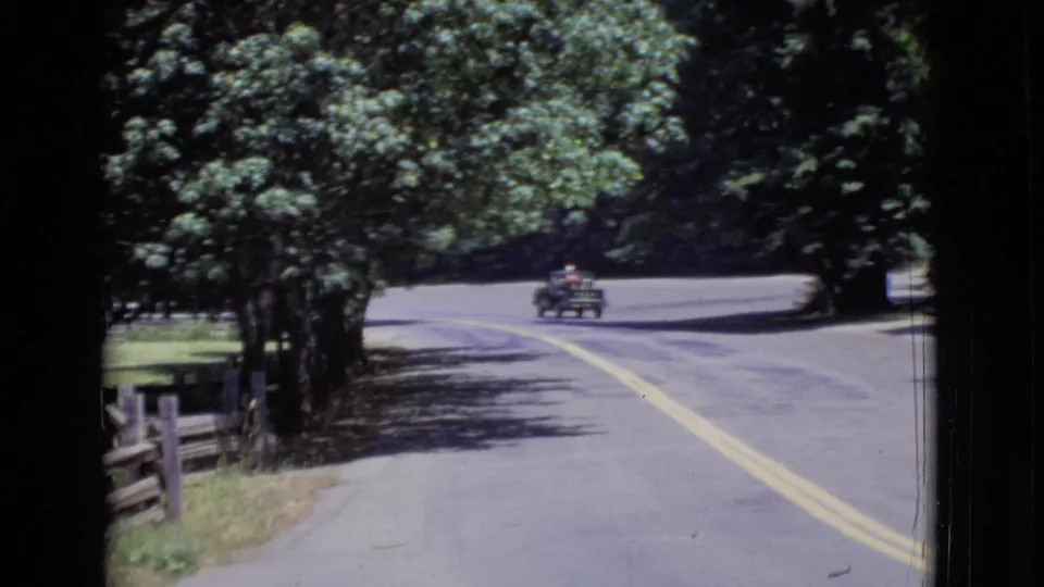 1968 San Diego County home-movie view of a tree-lined road, roadside fence, and small vintage car traveling through the wooded setting.
