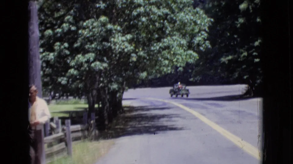 1968 San Diego County frame showing the man partly cropped near a wooden post, with trees, fence, and a small car on the road beyond.