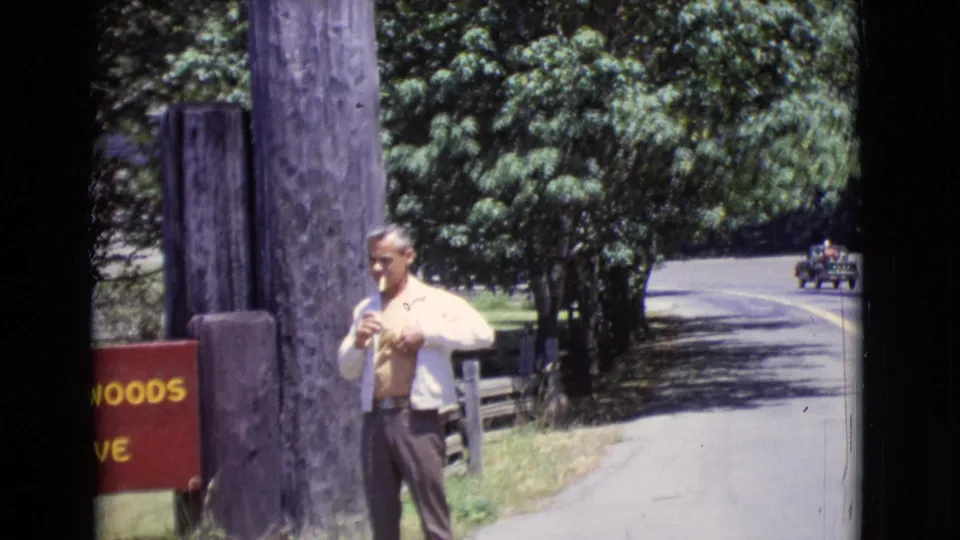 1968 San Diego County home-movie frame of a man beside the Redwoods Reserve entrance sign, holding a cigarette near the road.