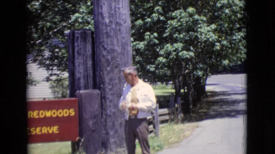 1968 San Diego County 8mm frame showing a man adjusting clothing near the Redwoods Reserve sign and roadside entrance posts.