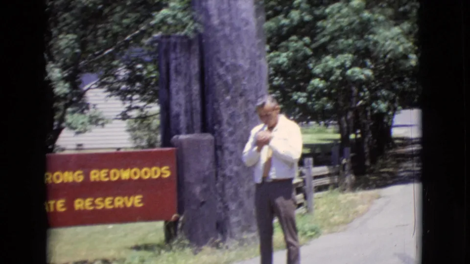 1968 home-movie frame of a man beside the Armstrong Redwoods State Reserve sign on a paved path, with trees behind him.