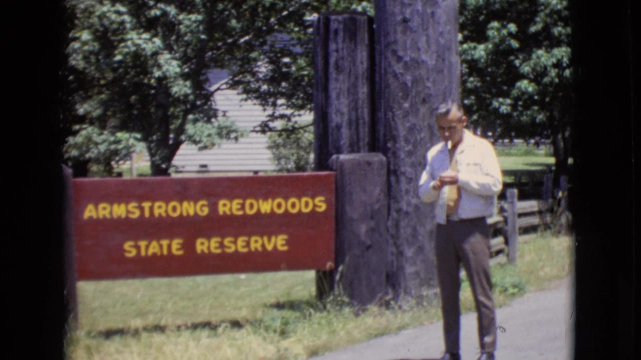A man in a light shirt and tie stands near the Armstrong Redwoods State Reserve sign, with trees and a path visible.