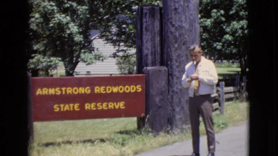 A man in a light shirt and tie stands near the Armstrong Redwoods State Reserve sign, with trees and a path visible.