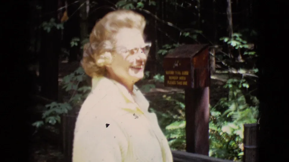 Woman in glasses smiles in the wooded reserve area, with a small posted sign and forest vegetation behind her.