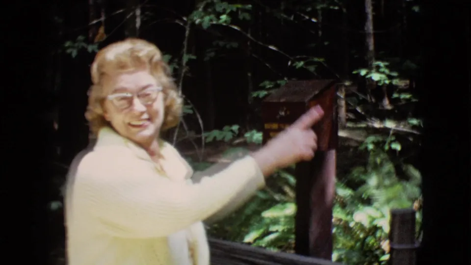 Woman in glasses points at a small posted sign in the wooded reserve area during the 1968 home-movie visit.