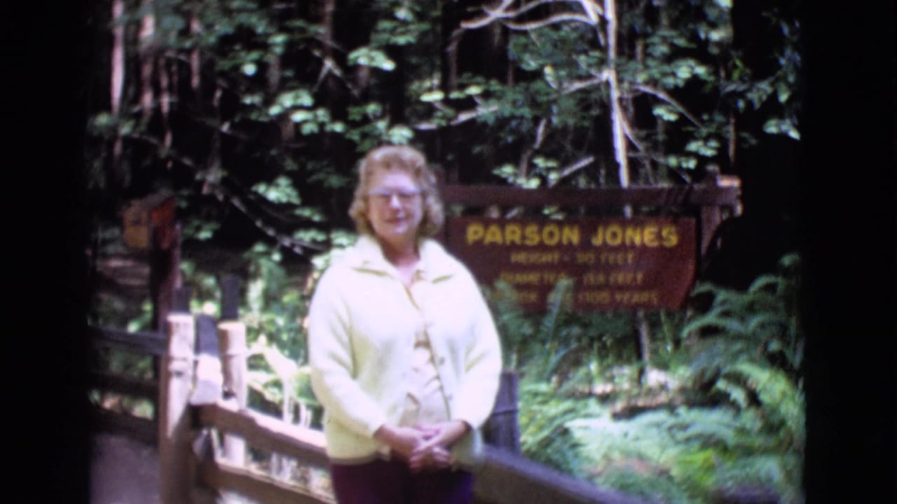 Woman standing beside a Parson Jones tree sign in a wooded area with a fence rail.