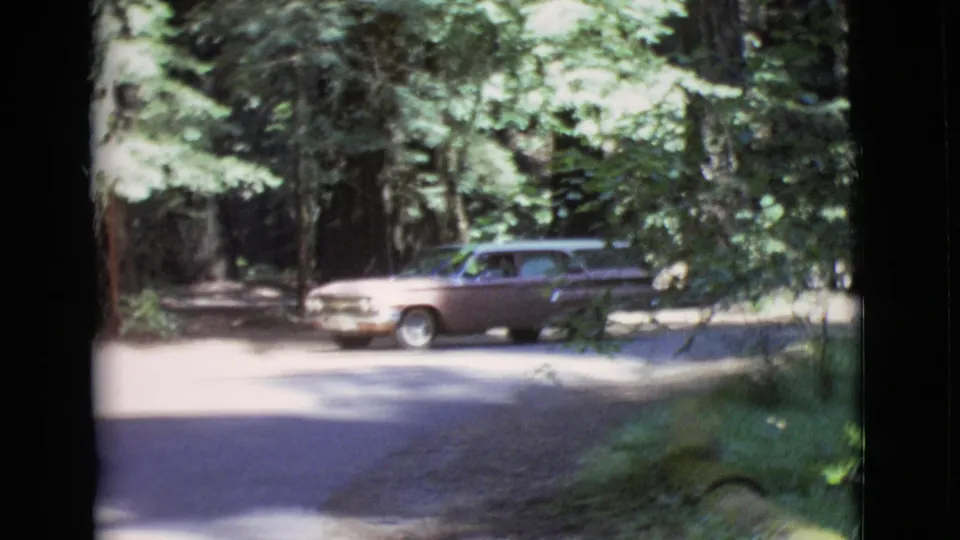 1968 San Diego County 8mm frame showing a brown station wagon passing across a bright forest road.