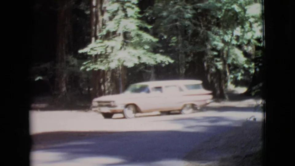 1968 San Diego County 8mm frame showing a light pink station wagon entering a sun-dappled forest road.