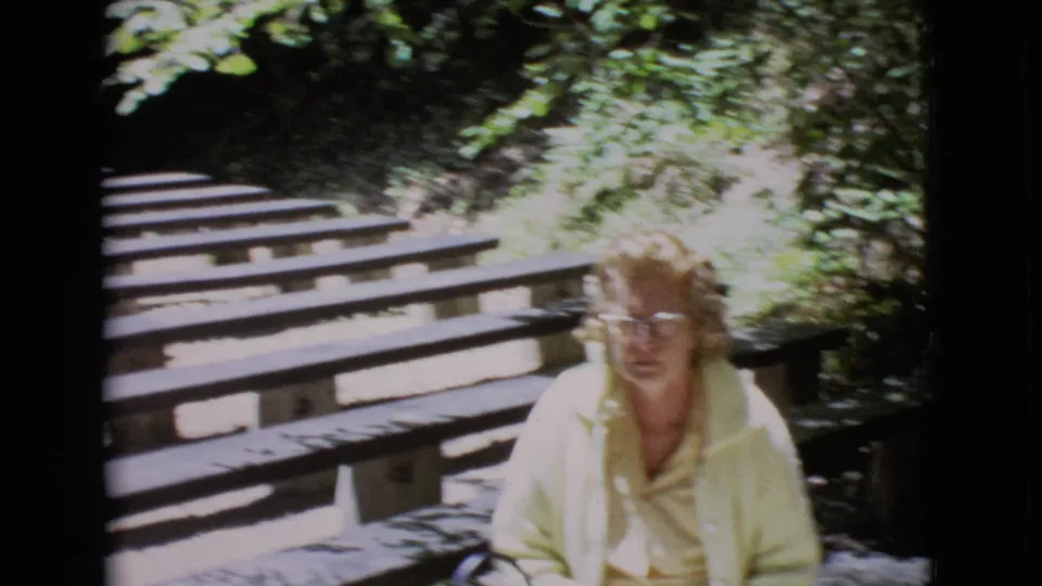 Adult woman in glasses and a yellow shirt sits beside wooden bleachers in a sunlit San Diego County outdoor setting.