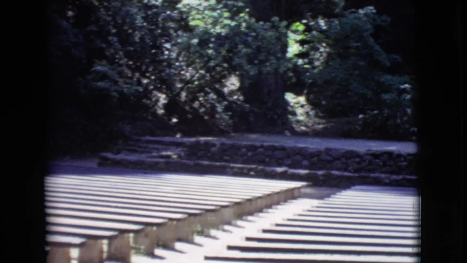 1968 San Diego County 8mm view across empty amphitheater seating, with bench supports, foliage, and a stone retaining wall visible.