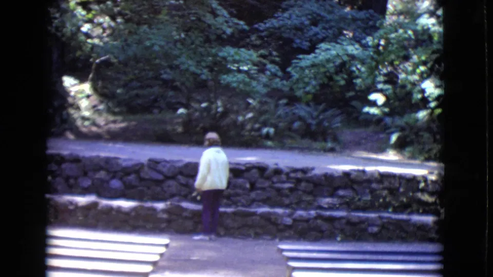 The 8mm frame shows a woman from behind on a paved path in a leafy San Diego County park, with benches and rockwork nearby.