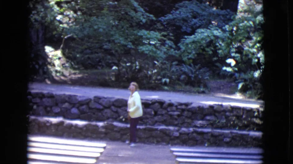 A woman turns partly toward the camera in 1968 San Diego County home-movie footage, standing near benches, rockwork, and shaded plants.
