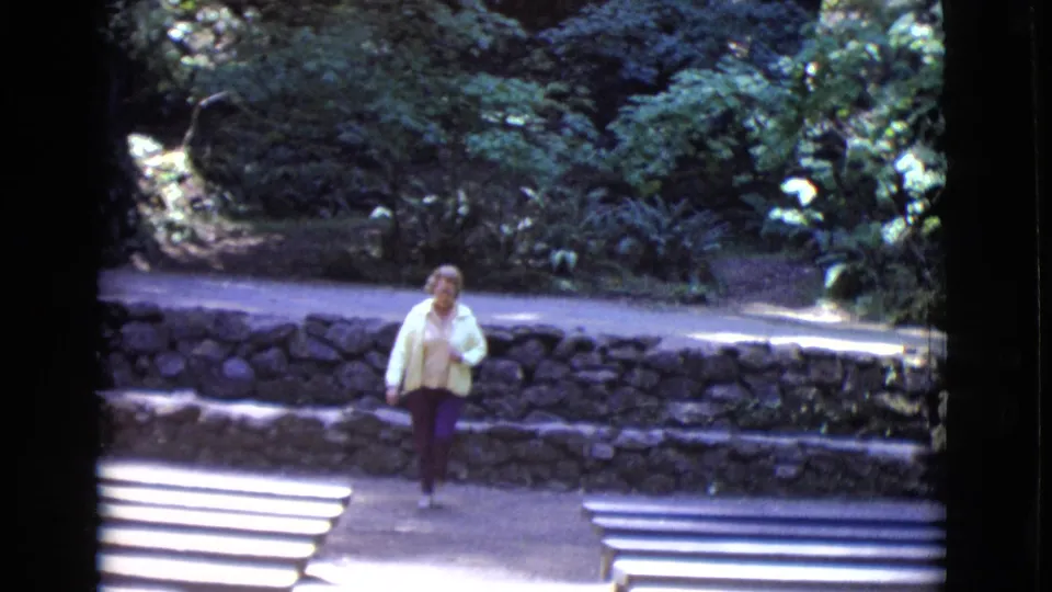 In 1968 San Diego County home-movie footage, a woman in a yellow jacket walks along a park path beside a stone wall and steps.