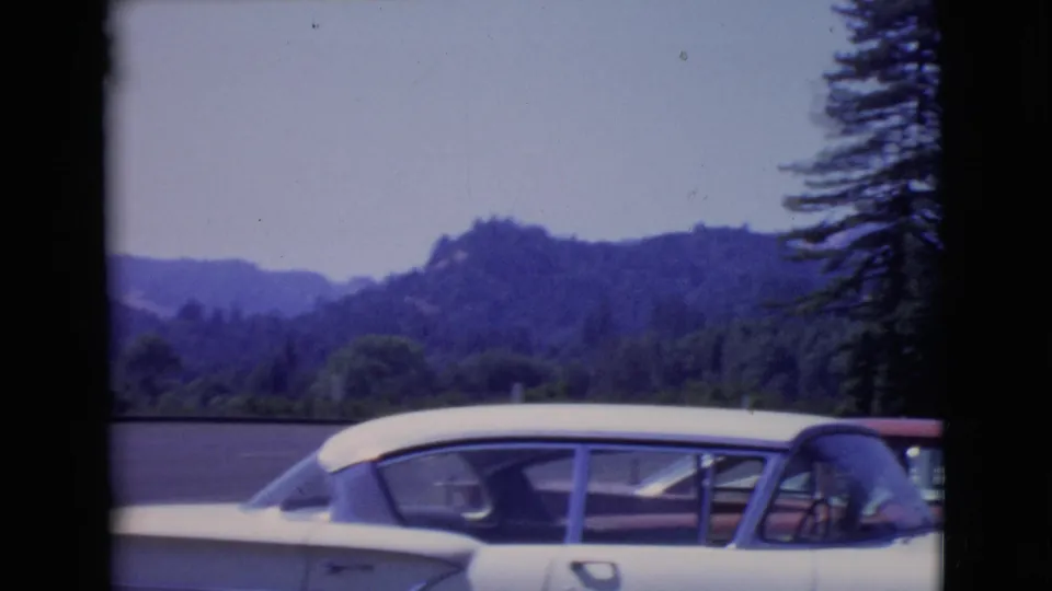 1968 San Diego County 8mm frame with a white 1960s car in the foreground, trees to the side, and wooded hills in the distance.