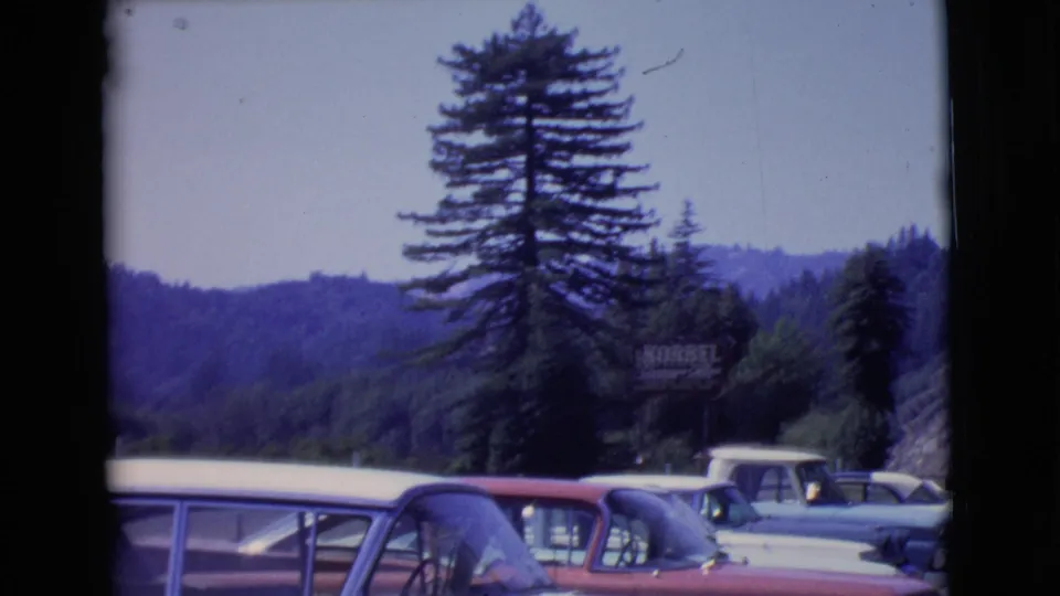 1968 San Diego County home-movie frame showing parked classic cars, forested hills, pine trees, and a KORBEL roadside sign.