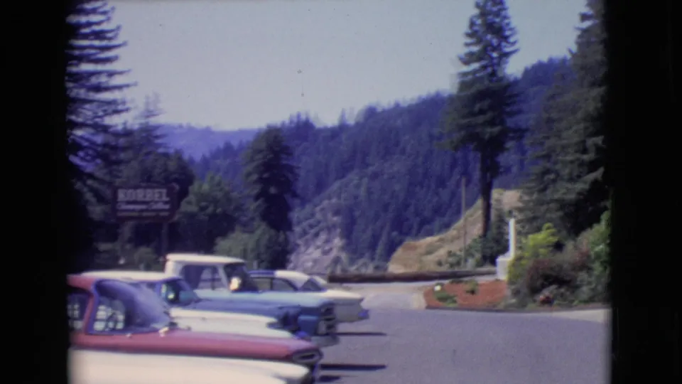 1968 San Diego County frame with classic cars beside a curving overlook road, wooded hills, a pine tree, and partial KORBEL signage.