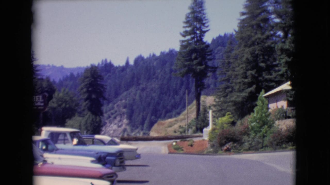 1968 San Diego County 8mm frame showing parked classic cars along a paved roadside near tall trees and a forested hillside.
