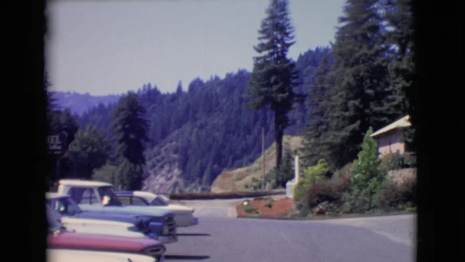 1968 San Diego County 8mm frame showing parked classic cars along a paved roadside near tall trees and a forested hillside.