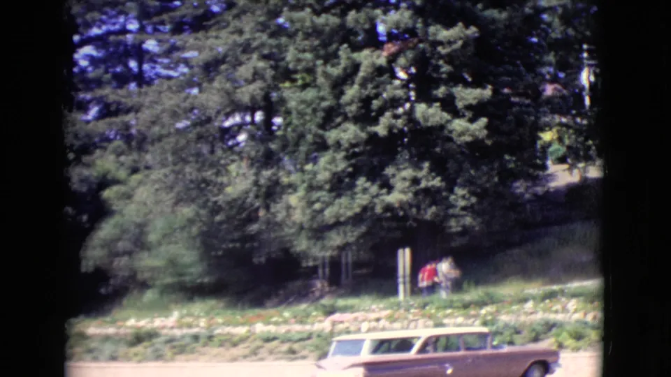 Two distant people walk near a planted hillside while a tan station wagon passes on the road in 1968 San Diego County footage.